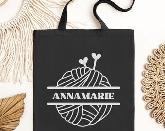 May include: Black tote bag with a white outline of a ball of yarn and knitting needles. The text "ANNAMARIE" is centered below the yarn.
