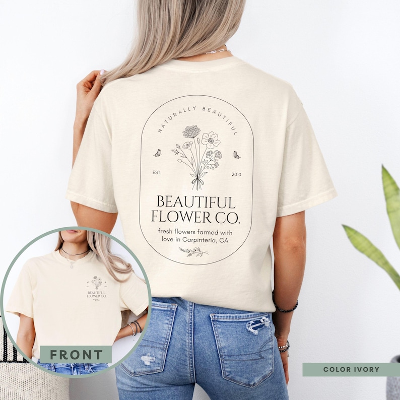May include: Ivory-colored t-shirt featuring a floral design with the text "BEAUTIFUL FLOWER CO." and "fresh flowers farmed with love in Carpinteria, CA." The design includes butterflies and the words "NATURALLY BEAUTIFUL."
