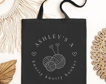 May include: Black tote bag with white text that reads "Ashley's a knotty knotty hooker" with a graphic of two yarn balls and knitting needles.