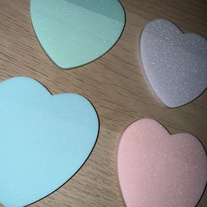 Könnte beinhalten: Vier herzförmige Kosmetikschwämmchen in Pastellfarben: Mintgrün, Hellblau, Lavendel und Rosa. Jeder Schwamm hat eine glitzernde, strukturierte Oberfläche, angeordnet auf einer Holzoberfläche.