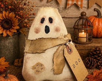 May include: A beige fabric ghost figurine with a hand-stitched appearance, featuring black painted eyes and an oval mouth. It wears a frayed burlap scarf and has a rectangular cardboard tag attached that is clearly printed with the word BOO.