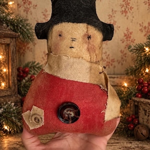 May include: A fabric snowman doll featuring a black top hat, a beige scarf, and a red body. The doll has embroidered star-shaped eyes and a stitched mouth. A dark brown button is centered on the red torso, with a smaller tan button on a patch.
