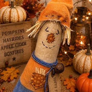 May include: A fabric scarecrow figurine featuring a tan burlap body, a pointed orange hat with frayed edges, and straw hair. It wears a blue base, has a stitched face, a small pocket with straw, a blue twine neck tie, and a tag reading HAPPY FALL.