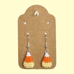 May include: Handmade crochet candy corn earrings. The earrings are white, orange, and yellow, with silver-coloured ear wires. They are displayed on a brown earring card. A fun accessory for Halloween.