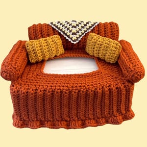 May include: A handmade crochet tissue box cover shaped like a miniature sofa. The cover is primarily rust-colored with mustard-yellow pillows and a small, patterned throw. The design is reminiscent of vintage decor.