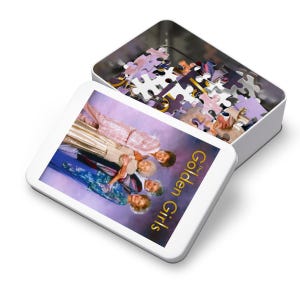 Può includere: Una scatola di latta bianca con un puzzle all'interno. La scatola ha un'immagine dei quattro personaggi principali dello show televisivo "The Golden Girls" sul davanti. I personaggi sono in piedi insieme e sorridono. Il testo "The Golden Girls" è scritto in lettere dorate sulla scatola.