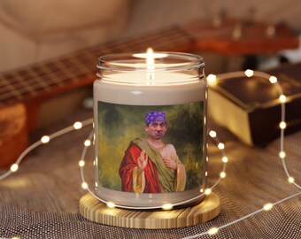 May include: A brown glass candle with a white wax top. The candle has a printed image of a man wearing a purple bandana and a red robe. The man is holding his hand up in a gesture of blessing. The candle is sitting on a wooden base.
