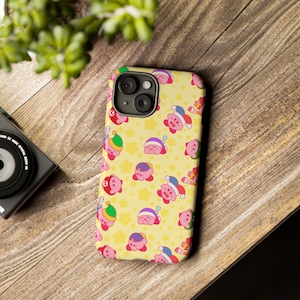Op de afbeelding: Een gele telefoonhoes met een herhalend patroon van roze Kirby-karakters die verschillende hoeden dragen. De hoes heeft een camera-uitsparing en een zwarte rand.