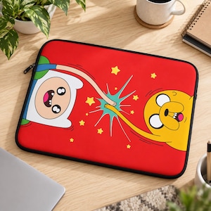May include: A rectangular laptop sleeve with a vibrant red background featuring a graphic illustration of Finn the Human and Jake the Dog high-fiving. The design includes yellow stars and a teal starburst effect between their hands.