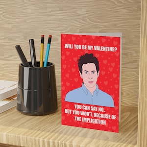May include: Valentine's Day greeting card with a red background and white text that reads "WILL YOU BE MY VALENTINE? YOU CAN SAY NO.. BUT YOU WON'T. BECAUSE OF THE IMPLICATION." The card features a cartoon illustration of a man in a blue shirt.