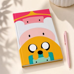 Può includere: Un taccuino con copertina rigida con un design a cartoni animati colorato, con una combinazione di colori rosa, bianco e giallo. C'è una penna rosa accanto al taccuino. La copertina presenta personaggi dei cartoni animati con grandi occhi e un volto sorridente.
