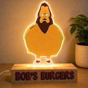 Könnte beinhalten: Eine beleuchtete Acryllampe mit einer Figur mit dunklem Bart in einem Burger-Kostüm, die auf einem rechteckigen Holzsockel steht, auf dessen Vorderseite der Text BOB'S BURGERS in fetten roten Buchstaben mit schwarzer Umrandung aufgedruckt ist.