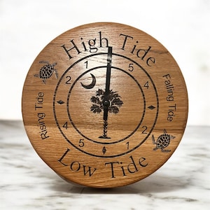 May include: A round wooden tide clock with engraved text and graphics. The clock face features the words "High Tide," "Low Tide," "Rising Tide," and "Falling Tide." A palm tree and crescent moon are in the center. Two sea turtles are on the clock face.