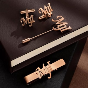 May include: A set of rose gold personalized accessories featuring the name "MJL" in a script font. The set includes cufflinks, a tie bar, and a lapel pin.