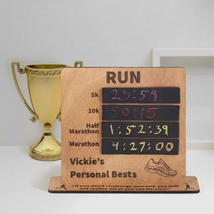 以下が含まれることがあります： 「RUN」と書かれた黒いチョークボードのインサートが付いた木製看板で、走行距離と書き込まれた時間がリストされています。距離は5km、10km、ハーフマラソン、マラソンです。時間は23:59、50:15、1:52:39、4:27:00です。看板には「Vickie's Personal Bests」とも書かれており、ランニングシューズのグラフィックが描かれています。看板には、「自分自身に挑戦しなければ、自分が何者になれるのか気づかないだろう」という引用文が書かれています。
