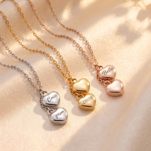 Könnte beinhalten: Drei zarte Halsketten mit dünnen Metallketten, jede mit zwei kleinen herzförmigen Anhängern. Die silberne Kette ist mit 'Coco' und 'Mom' graviert, die goldene mit '0908' und die roségoldene Halskette trägt die Gravur 'Bella'.