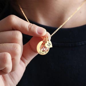 Puede incluir: Collar dorado con un colgante con el nombre "MACK" y un dije en forma de corazón con la letra "M". La cadena es delicada. El collar está sostenido por una mano.