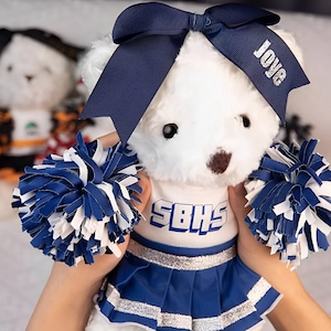 May include: White plush teddy bear wearing a blue and silver cheerleader outfit with pom-poms. The bear has a blue bow with the word "LOVE" on it and a white shirt with the text "SBHS".
