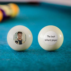 May include: Two white billiard balls on a teal pool table. One ball has a photo of a smiling person, the other has the text "The best billiard player" on a white circle. The balls are for a billiards theme.