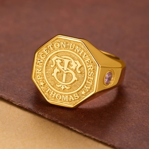 May include: A gold-colored ring with an octagonal face. The face features the words "PRINCETON UNIVERSITY THOMAS" around a central emblem. A small, round, purple stone is set into the side of the ring.