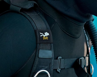 Könnte beinhalten: Ein schwarzes Tarierjacket zum Tauchen mit einem gepolsterten Schultergurt, der eine weiße Taucher-Silhouette und den gelben Schriftzug Diver zeigt. Die Ausrüstung umfasst verstellbare Nylongurte, Kunststoff-Schnallen und einen Metall-D-Ring.