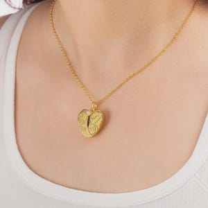 May include: A gold-colored necklace with a heart-shaped pendant. The pendant features a pair of angel wings and a small, clear gemstone. The necklace is worn around the neck.