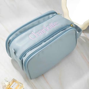 May include: A pale blue cosmetic bag with the name "Charlotte" embroidered in pink. The bag features multiple zipped compartments and a small handle. The bag is made of a smooth, leather-like material.
