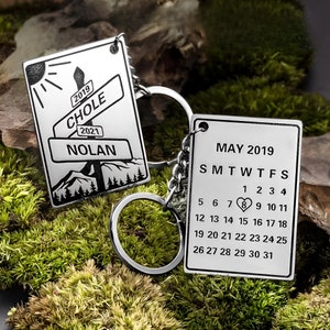 Könnte beinhalten: Silberner Schlüsselanhänger mit zwei Anhängern. Der erste Anhänger zeigt eine Bergszene mit dem Text "2019 CHOLE 2021 NOLAN". Der zweite Anhänger ist ein Kalender für Mai 2019 mit einem Herz, das den 8. markiert.