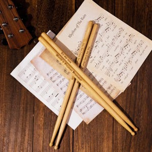 Peut inclure: Baguettes de batterie en bois avec texte gravé, posées sur une partition et une surface en bois. Les baguettes portent les inscriptions "To my son, World's Greatest Drummer" et "You Are Every Beat Of". La partition est intitulée "Rhythm of The Rain".