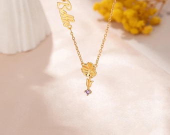 May include: A gold-colored necklace with the name "Bella" in cursive script. The necklace features a flower pendant with a purple gemstone and a tulip-shaped element. The chain is delicate, and the background is a soft pink.