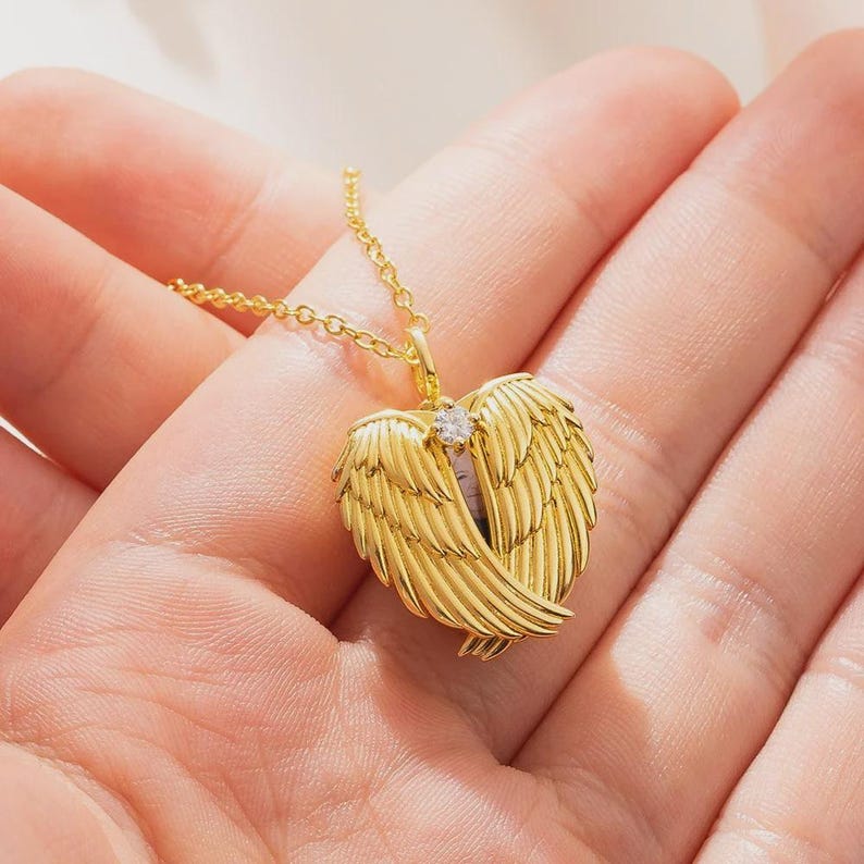 May include: A gold-colored necklace featuring a heart-shaped pendant with angel wings. The pendant has a small, clear gemstone in the center. The necklace is on a delicate chain, and the wings are detailed with fine lines.