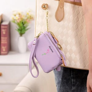 May include: A lavender-colored wallet with a wrist strap and a gold-tone zipper. The wallet is attached to a handbag with a gold chain. The word "Florence" is printed on the wallet, along with a floral design.