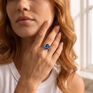 May include: A silver ring featuring an oval-shaped, iridescent blue and purple mood stone set in a polished metal band. The ring is worn on the ring finger, showcasing the smooth, reflective surface of the cabochon stone and the metallic setting.