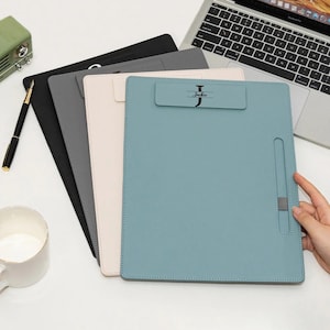 May include: A stack of four leather clipboards in black, gray, cream, and teal. The teal clipboard has the name "Jackie" printed on it. A pen and a mug are visible in the background.