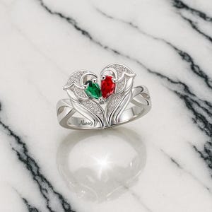 Puede incluir: Un anillo plateado con un dise&ntilde;o en forma de coraz&oacute;n, con una gema verde y otra roja. El anillo est&aacute; adornado con peque&ntilde;as piedras transparentes y la palabra "Always" est&aacute; grabada en la banda. El anillo est&aacute; sobre un fondo de m&aacute;rmol.