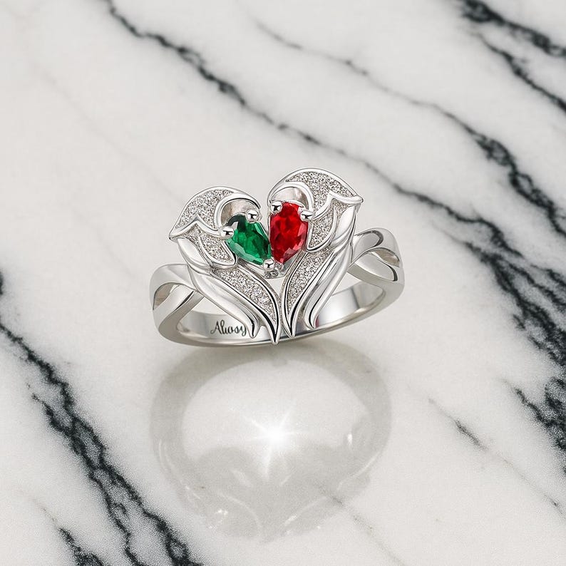 Puede incluir: Un anillo plateado con un dise&ntilde;o en forma de coraz&oacute;n, con una gema verde y otra roja. El anillo est&aacute; adornado con peque&ntilde;as piedras transparentes y la palabra "Always" est&aacute; grabada en la banda. El anillo est&aacute; sobre un fondo de m&aacute;rmol.
