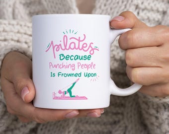 May include: A white ceramic mug featuring pink and green text that reads Pilates Because Punching People Is Frowned Upon, accompanied by a graphic of a person performing a pilates exercise on a machine with pink and green accents.