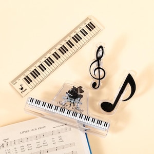 May include: A collection of music-themed stationery items. Includes a ruler with a piano key design, a treble clef, a quarter note, and a clip with a piano key design and the text "Sadie's Music".