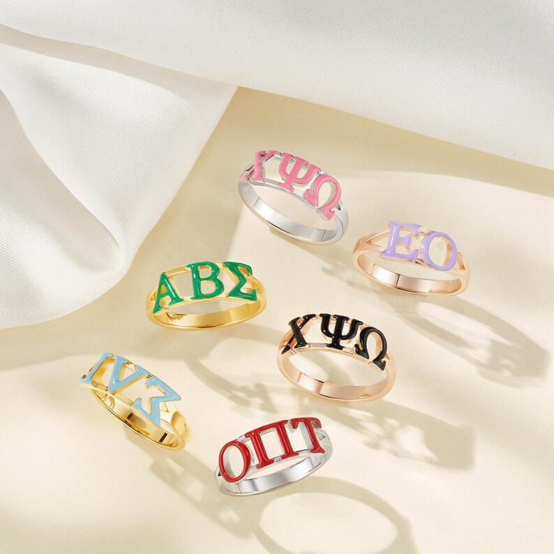 May include: A collection of colorful enamel rings, each featuring Greek letters in various colors. The rings are gold and silver-toned, with letters in green, pink, purple, black, red, and light blue. The rings are displayed on a light-colored surface.