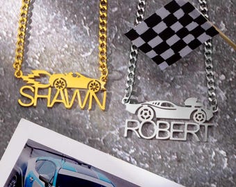 May include: Two necklaces featuring car designs. One is gold-coloured with the name "SHAWN" and a car silhouette with flames. The other is silver-coloured with the name "ROBERT" and a car silhouette. A checkered flag is also present.