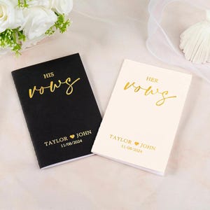 Puede incluir: Dos libros de votos de boda, uno negro y otro crema. El libro negro dice "HIS vows" en letras doradas, con los nombres "TAYLOR JOHN" y la fecha "11/08/2024" debajo. El libro crema dice "HER vows" en letras doradas.