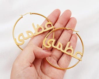 May include: A pair of gold hoop earrings with the Arabic word "sabba" written on them.
