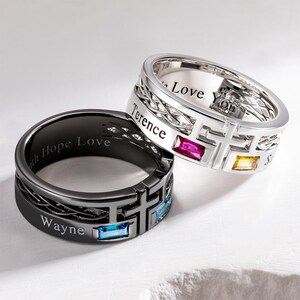 May include: Two metal rings featuring a cross design and baguette-cut gemstones. The silver-toned ring is engraved with "Terence" and "Love You" with red and yellow stones. The black ring is engraved with "Wayne" and "Faith Hope Love" with blue stones.