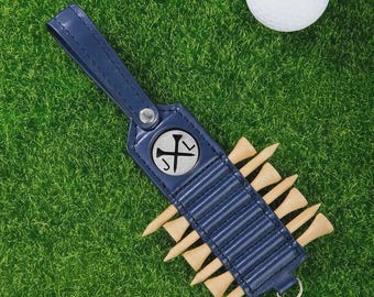 May include: A navy blue leather golf tee holder with a silver emblem and wooden tees, resting on green grass next to a golf ball. The holder has a loop for attachment.
