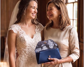 May include: A navy blue satin clutch bag with a silver chain strap and a magnetic snap closure. The interior flap features a black and white printed photograph of a person and a child. The clutch is held by a person in a champagne dress.