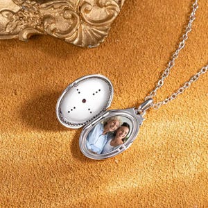 May include: Silver oval locket necklace with a photo of two people inside. The locket has a small red stone on the front.