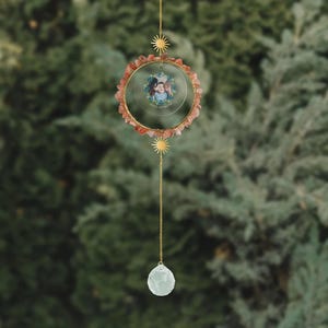 May include: A hanging suncatcher with a gold-colored metal frame. The frame is adorned with small, reddish-orange stones and a small photo. A clear crystal ball hangs from a chain below, and a sun-shaped charm is above.