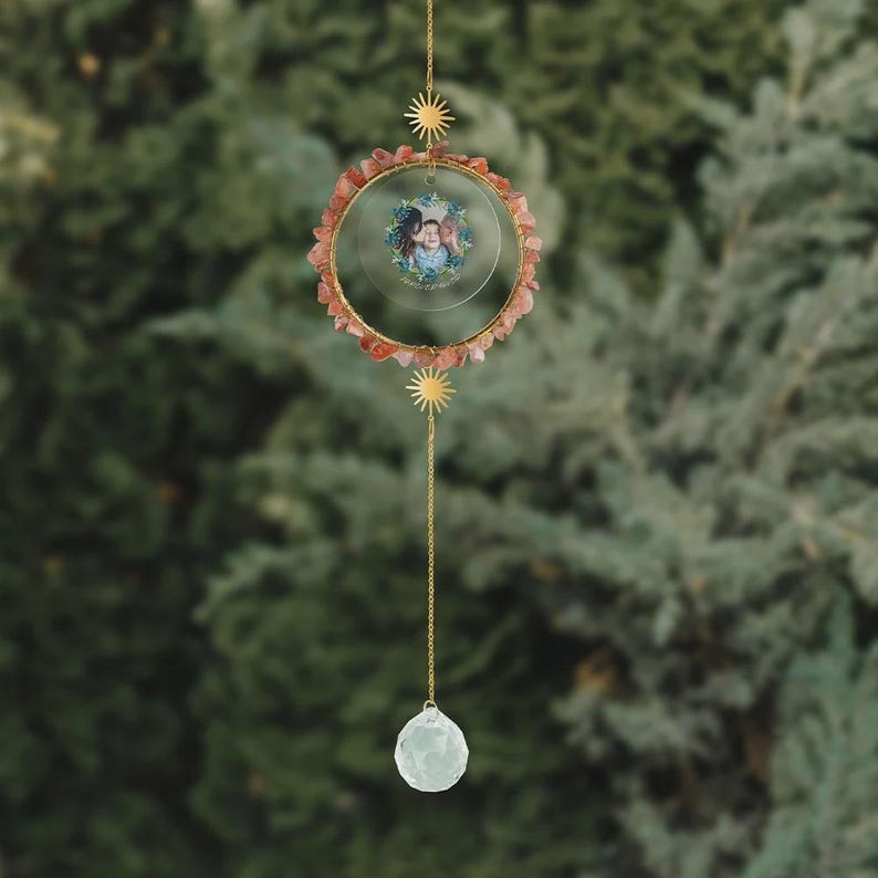 May include: A hanging suncatcher with a gold-colored metal frame. The frame is adorned with small, reddish-orange stones and a small photo. A clear crystal ball hangs from a chain below, and a sun-shaped charm is above.