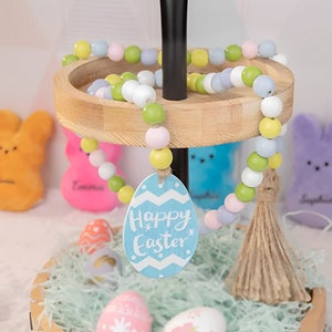 以下が含まれることがあります： 緑、黄色、ピンク、ブルーの色合いの木製ビーズでできたパステルカラーのイースターガーランドがついた、段になった木製スタンド。ガーランドには、「Happy Easter」という文字が書かれた青い木製卵がぶら下がっています。スタンドは、パステルカラーのイースターエッグと青と白のイースターバニーで飾られています。