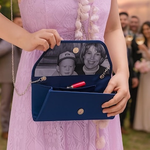 K&ouml;nnte beinhalten: Eine marineblaue Clutch mit silberner Kette, mit einem Schwarz-Wei&szlig;-Foto im Inneren. Ein roter Lippenstift ist im offenen Fach sichtbar. Die Clutch wird von einer Person in einem lavendelfarbenen Kleid gehalten.